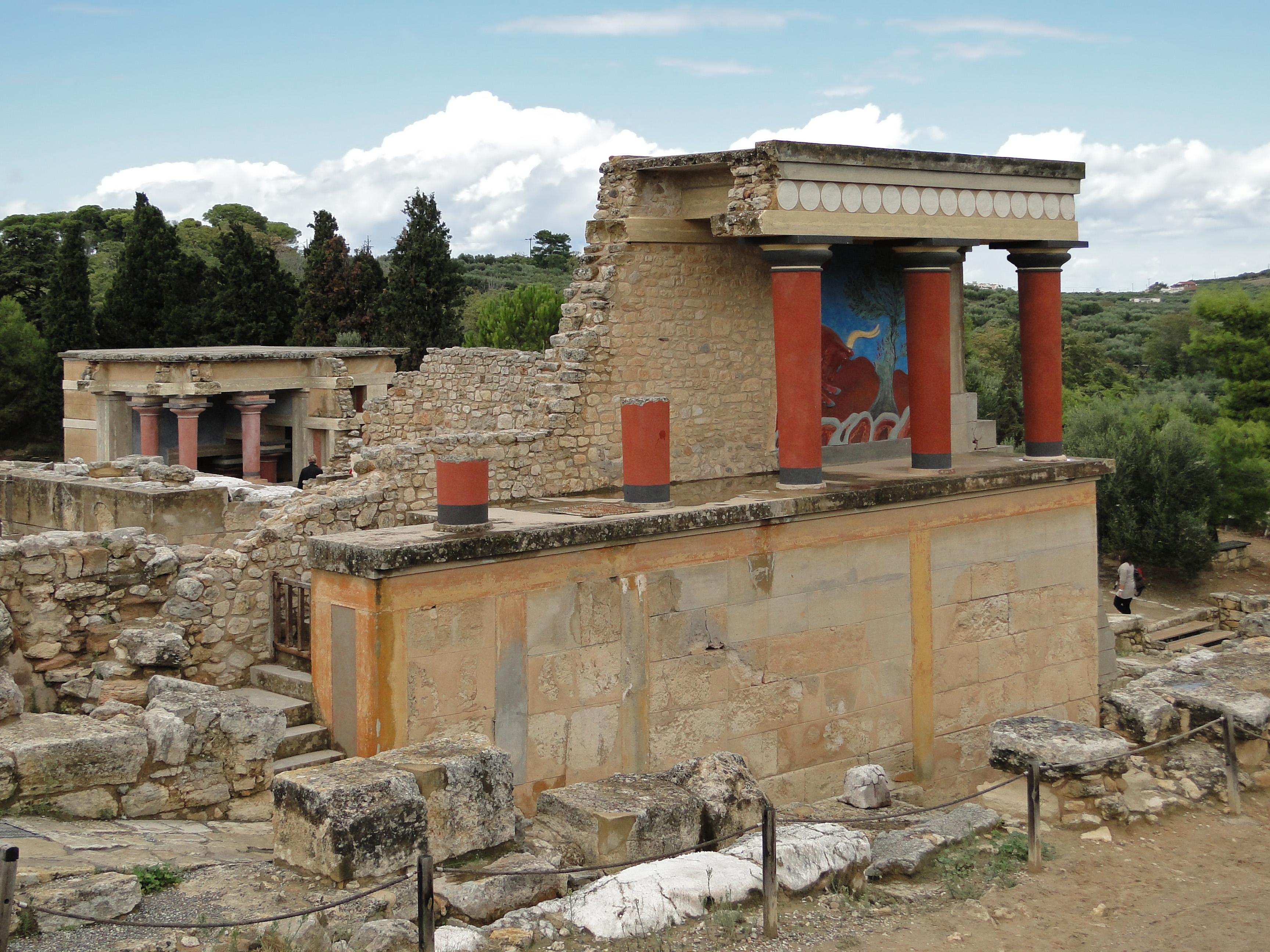 An image of King Minos in royal dress seated before the palace at Knossos with Cretan columns and sea trade ships in the background.